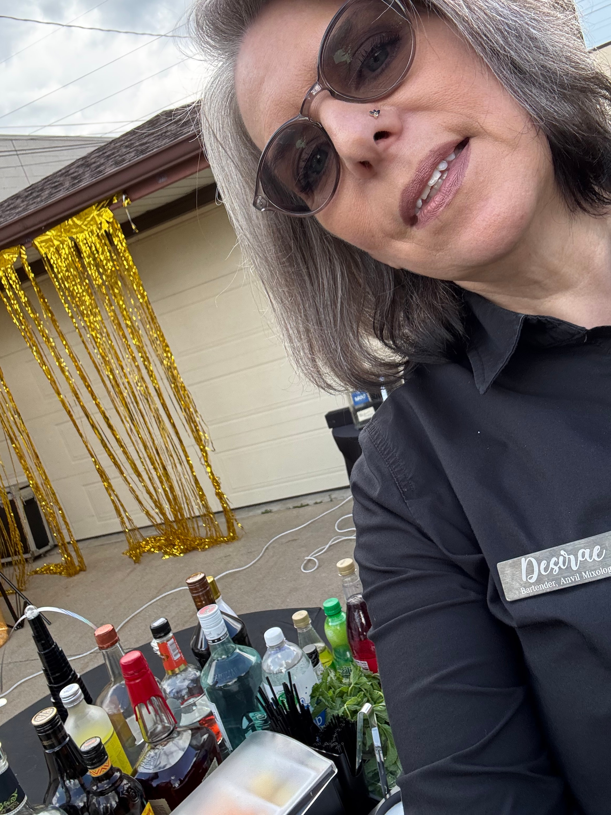 Desirae smiling at the bar with gold fringe backdrop and full bottle lineup at a housewarming event