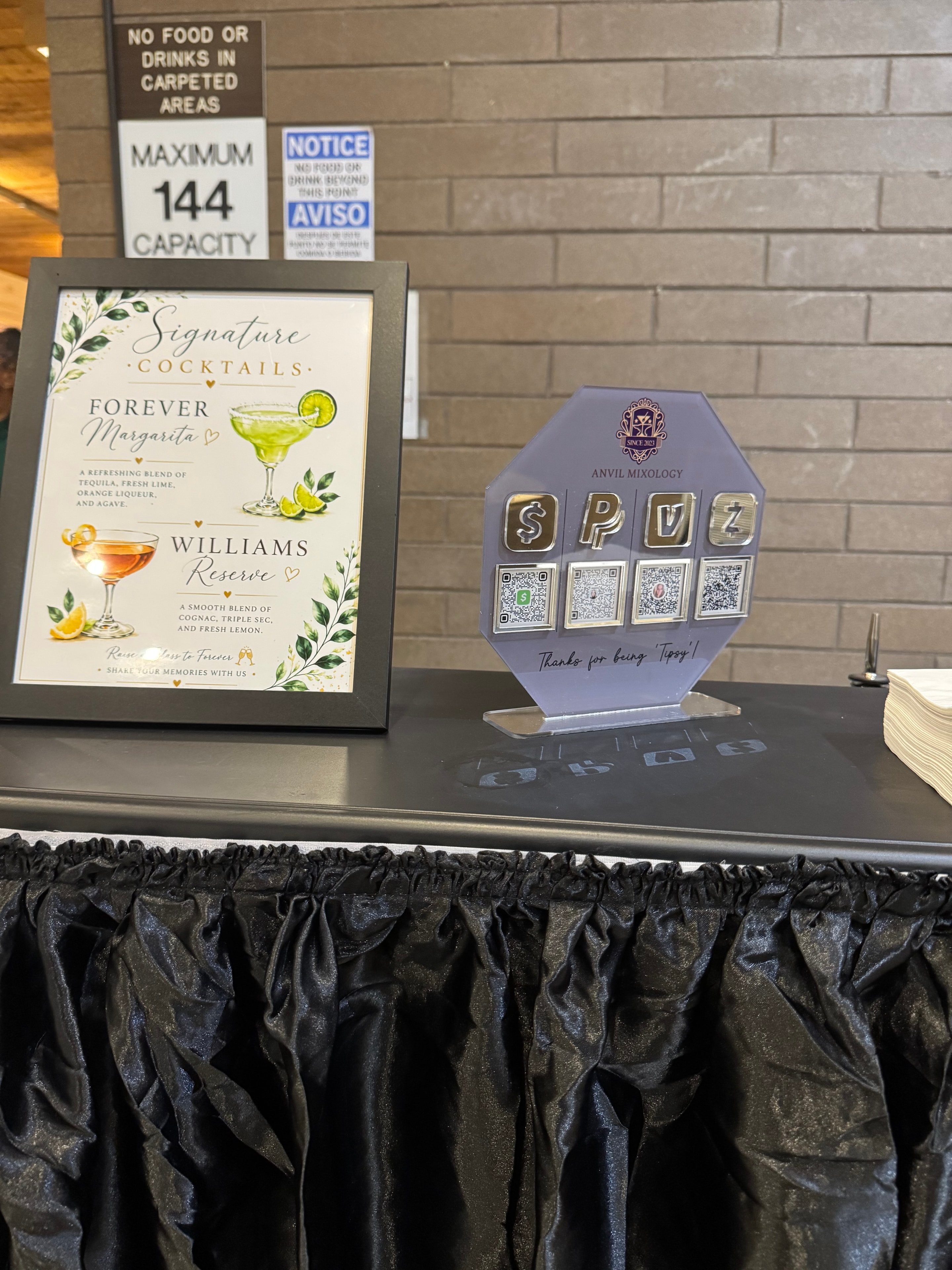 Close-up of the Anvil Mixology bar station with signature cocktail menu, QR code tip display, and custom napkins at the Bryan & Kristiane wedding