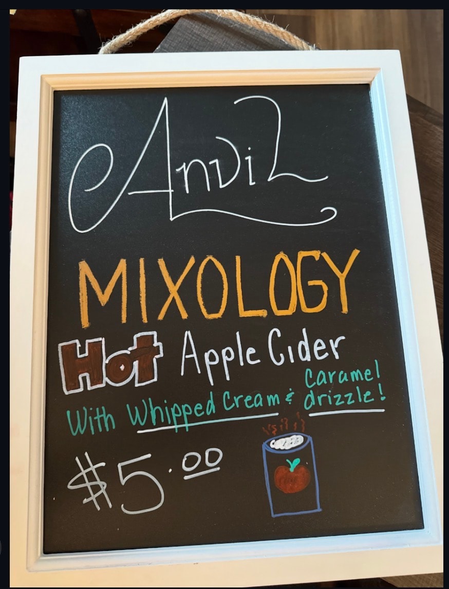 Anvil Mixology chalkboard sign for Hot Apple Cider with whipped cream and caramel drizzle at a winter gathering